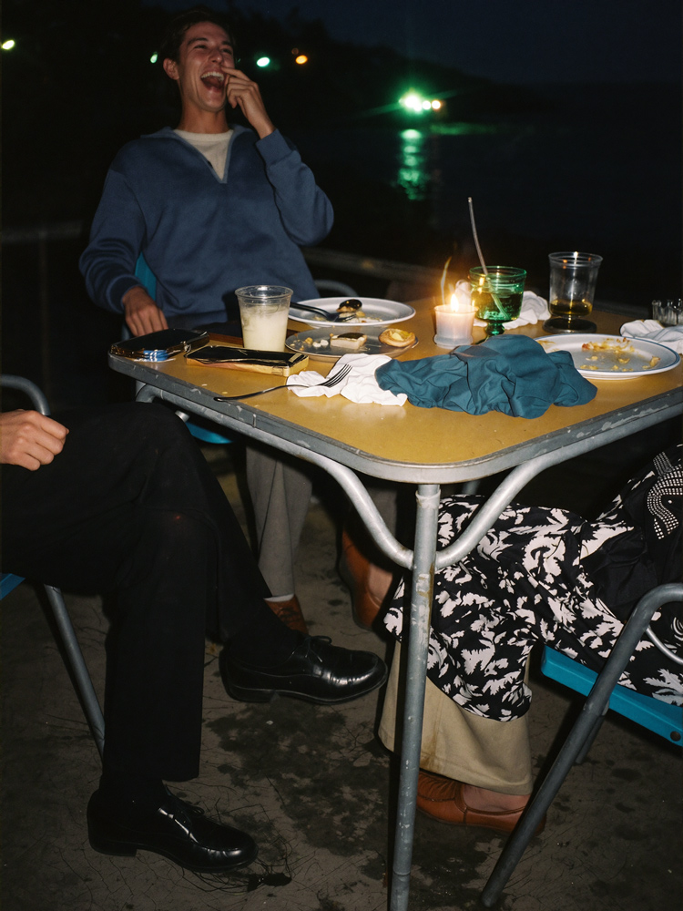 Un hombre que se ríe, llevándose la mano a la boca, en una mesa al aire libre iluminada con velas por la noche, rodeado de platos y bebidas.