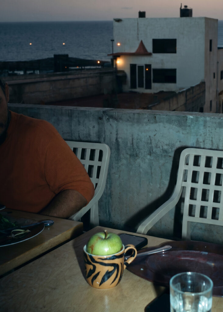 Una persona con una camiseta naranja sentada en una mesa al aire libre en una azotea o terraza, con una taza en la que hay una manzana verde, un plato y unos vasos, mientras que, al fondo, se divisan el mar y los edificios en el crepúsculo.