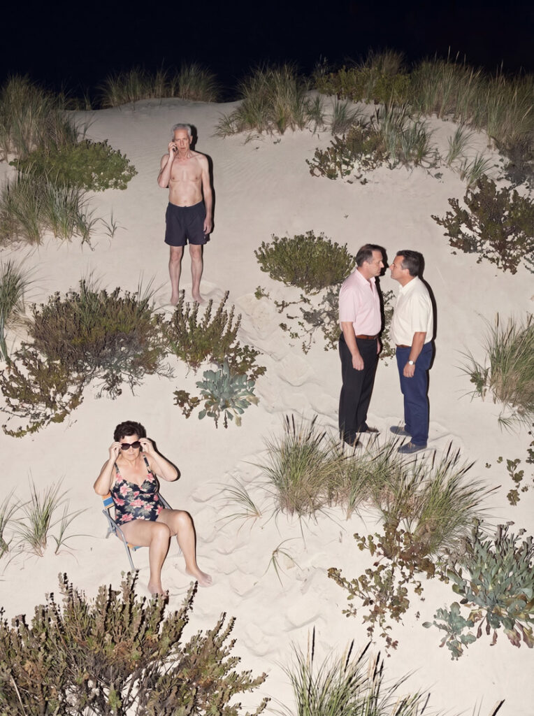 Escena nocturna en las dunas con tres personas: dos hombres besándose, una mujer con un bañador de flores que se ajusta las gafas de sol sentada en una silla, y un hombre sin camiseta en una duna hablando por teléfono.
