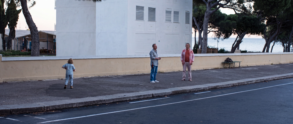 Un niño está solo en una acera, junto a un muro bajo, mientras dos adultos conversan cerca de él, con los árboles y el mar al fondo.