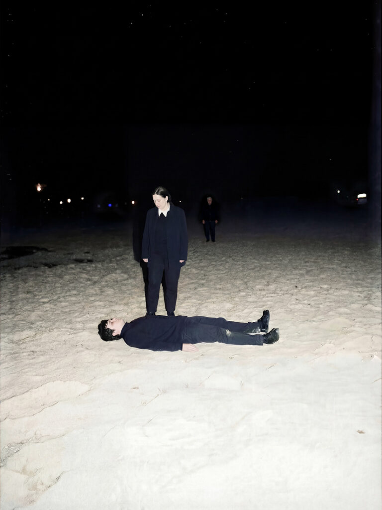 Escena nocturna en la playa: una persona tumbada en la arena mientras otra permanece de pie a su lado, con un cielo oscuro y figuras lejanas al fondo.