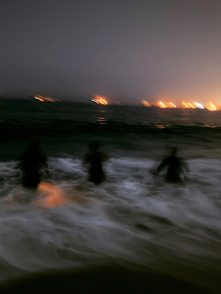 Escena nocturna borrosa en la que se ven tres siluetas vadeando entre las olas embravecidas de la orilla, con las luces de la ciudad en el horizonte y un cielo sin luna.
