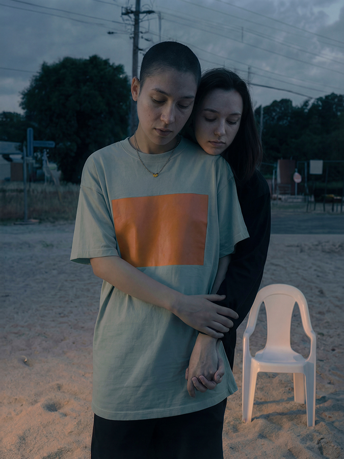 Dos personas en una playa de arena al atardecer: una abraza a la otra por la espalda, ambas con los ojos cerrados, junto a una silla de plástico blanca y con las tenues luces de la calle al fondo.