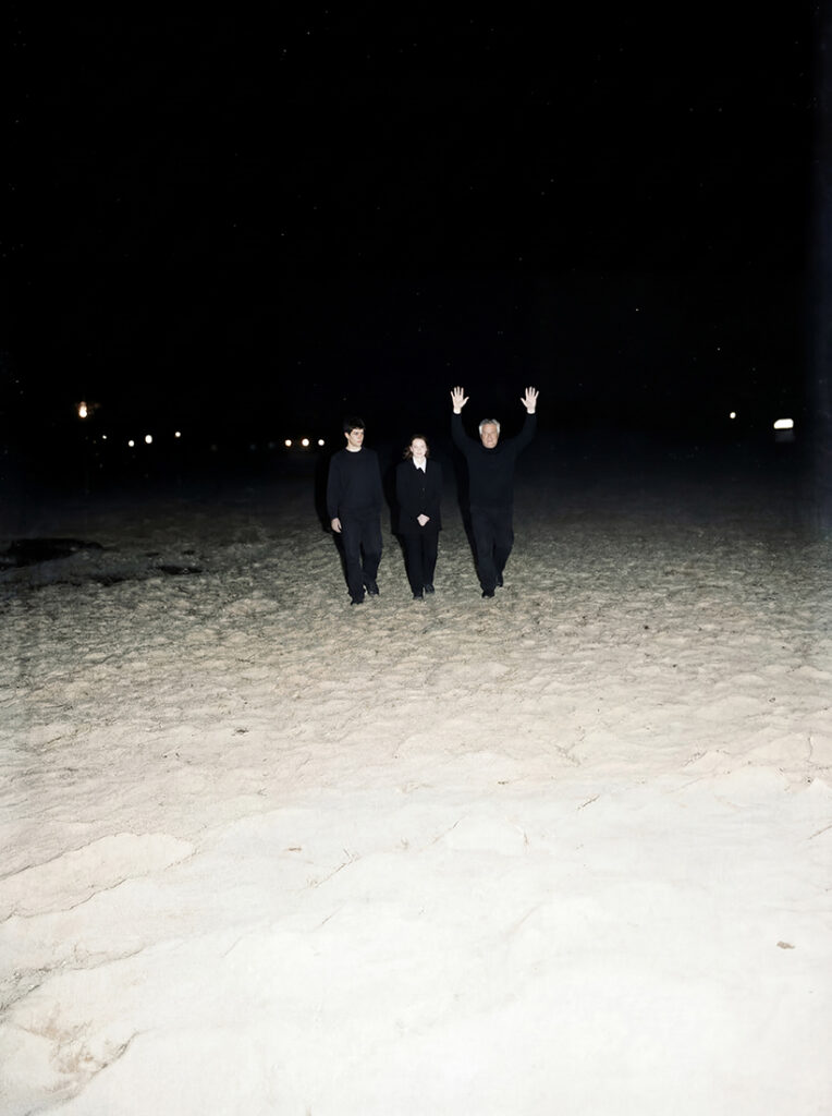 Cuatro personas vestidas de negro caminan hacia la cámara por una playa de arena por la noche; dos llevan los brazos a los lados, una levanta ambos brazos en el aire, con luces lejanas en el horizonte.