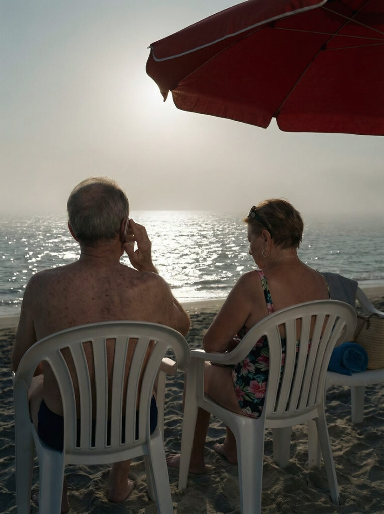 Una pareja sentada en la playa en unas sillas de plástico blancas bajo una sombrilla roja, mirando hacia el mar al atardecer; el hombre habla por teléfono mientras la mujer descansa a su lado.