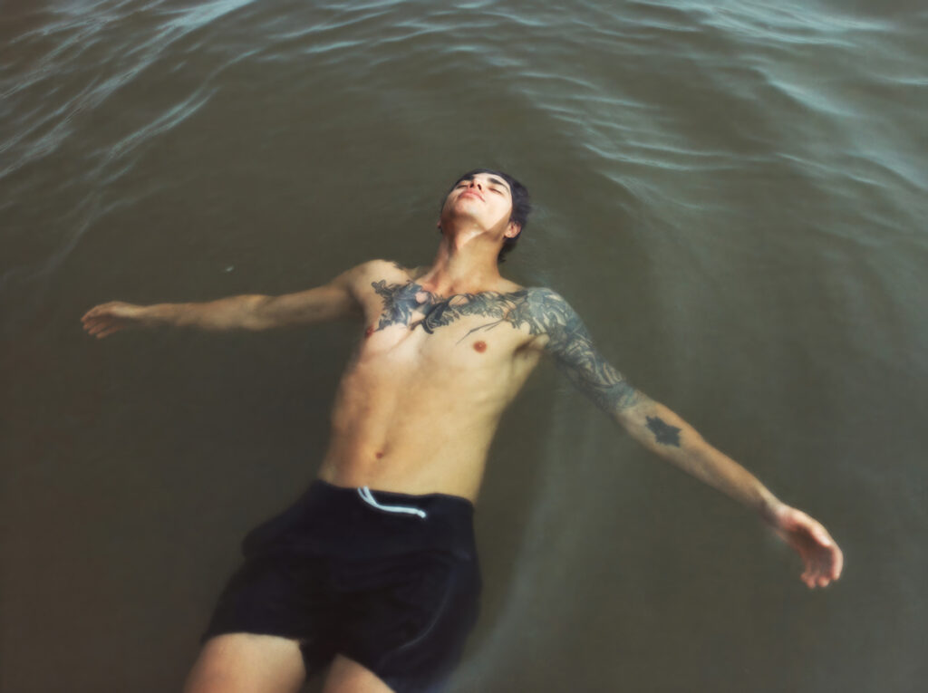 Un hombre sin camiseta flotando boca arriba en el agua, con los brazos abiertos y los tatuajes del pecho y los brazos a la vista.