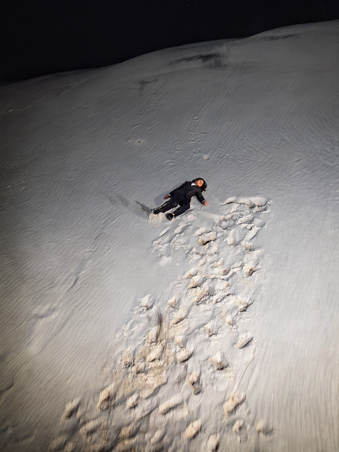 Person lying on a snow-covered slope at night, with footprints trailing down the hill.