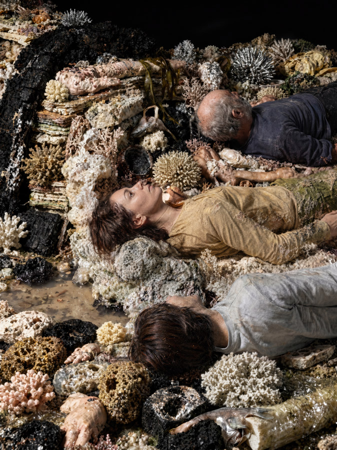 Three people lie resting among a colorful coral reef and rocks, appearing exhausted or posing on the reef-covered surface.