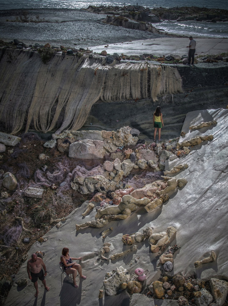 People explore a rocky coastal area with layered, bone-colored formations along a hillside and near a sand/stone shore. A child in a green outfit stands among the rocks while two adults sit nearby and a man looks over the edge from the top.