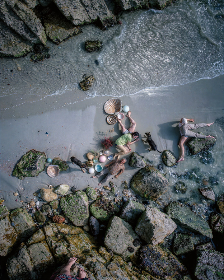 Aerial view of people sifting shells and sea glass on a sandy, rocky beach by the waves.
